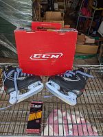 Pair of black CCM youth ice hockey skates on a metal rack, with original red CCM box behind them and a box of hockey blade wax in front.