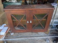 Full view of antique primitive wooden hanging cupboard with glass-front double doors, showing crisscross wooden trim and two glass doorknobs.