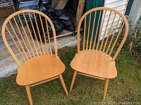 Two oak Windsor chairs shown upright on grass outside, displaying their curved spindle backs and contoured seats.