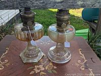 Two Aladdin Nu-Type Model B lamp bases with cathedral and beehive glass patterns, metal burners with wick adjusters, set on a decorated table outdoors.