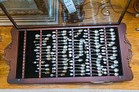 Wooden wall-mounted glass front display cabinet showing organized rows of silver plated collector spoons on black felt background.