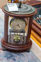 Bombay footed mantel clock with wood and metal construction, ornate top and feet, glass front and sides. Roman numeral clock face visible.