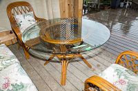 Photo showing full round beveled glass top on rattan base with floral cushioned chairs around it on a wooden deck.