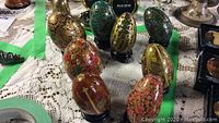 Eight hand painted wooden eggs with various floral and abstract designs, some with noticeable paint chips. Eggs are displayed on black stands on a lace cloth with other antiques in background.