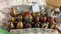 Photo showing an egg carton holding 15 hand painted wooden eggs with diverse intricate floral and ornamental patterns, predominantly gold, red, and black, some with wear and chips.