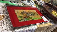 Photo showing hand-painted metal and glass tray with red border and horse carriage rural scene illustration on cardboard backing.