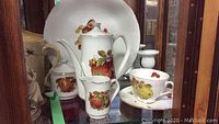 Photo showing teapot, creamer, sugar bowl, teacup, saucer, and large plate base with fruit pattern on white porcelain.