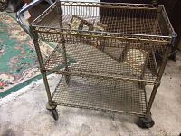 Metal wire rack with 3 wire mesh shelves and wheels on concrete floor. Shows rust and wear on metal frame and shelves.