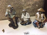Three mud figurines of elderly Chinese men in traditional clothing, posed differently, displayed on a shelf.