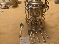 Front view of silver plated sugar bowl surrounded by multiple silver spoons on a table with yellow tablecloth.