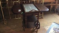 Full view of the wooden tea cart showing two drop leaves extended with a removable glass tray in the middle and large wooden wheels on one side.