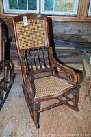 Front view of the antique wooden rocking chair showing cane mesh backrest with spindle details and scrolled armrest on wooden rocking base.
