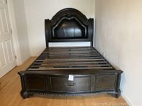 Queen size platform bed with padded black leather or faux leather upholstered headboard and dark wood frame with carved details and nailhead trim.