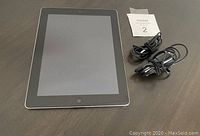 Apple iPad 2 laying face up with two black charging cords beside it on dark wooden surface.