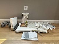 Photo of all lot contents on the floor against a wall showing Nintendo Wii console, Wii Fit balance board, Wii remotes and attachments, and multiple game cases stacked vertically.