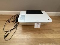 White HP Deskjet 1513 printer with power and USB cables attached, sitting on a wooden floor.