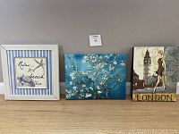 Photo of three paintings: beach-themed framed print, floral blossom canvas painting, and London art with Big Ben and woman