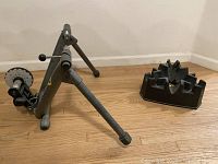 Metal stationary bike stand with adjustable lever and rubber feet next to a black front wheel riser block on wooden floor.
