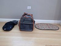 Overview of airline pet carrier, black pet jacket, and food bowl mat on floor.