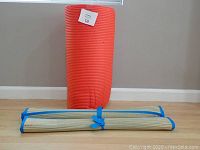 Red ribbed yoga mat standing vertically with a beige yoga mat rolled and tied horizontally in front, both on a wooden floor against a beige wall.