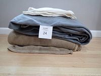 Three stacked fleece blankets in gray, beige, and cream on a hardwood floor against a gray wall.