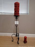 Large and small red carved candles with black metal stand indoors on wooden floor near window with blinds.