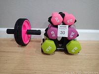 Photo shows pink 2 lb, purple 3 lb, and green 5 lb dumbbells on black AmazonBasics rack with ab wheel to left.