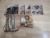 Overview photo showing tiered wooden box with multiple compartments containing costume necklaces, bracelets, rings, and three wristwatches placed on a wooden floor.