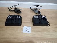 Photo of two remote control helicopters and two black remote controllers placed on a wooden floor with a lot number card in the middle.