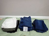 Photo of 8 pairs of jeans folded and stacked on a green surface against a wall, showing different colors and styles with a MaxSold card marked 49.