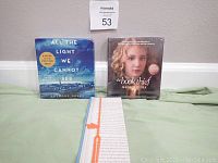 Two boxed audiobook sets and a bookmark laid out on a green surface, showing the front covers of both books.