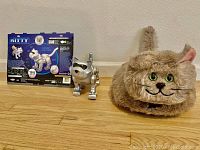 The photo shows two cat toys positioned side by side on a wooden floor against a beige wall: a silver metallic robot cat toy with black eyes and nose, and a plush fluffy cat toy with big green eyes, pink ears, and whiskers.