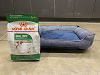 Photo showing a sealed bag of Royal Canin Small Breed Adult dog food next to a blue dog bed with tags.