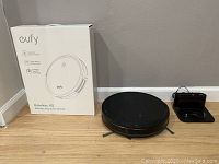 Eufy RoboVac 11S robotic vacuum cleaner, original box, and charging station arranged together on a wooden floor against a wall.