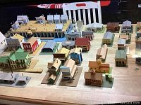 Wide view of multiple balsa wood buildings showing different styles and sizes on a wooden surface with a chair in background.