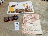 Photo showing a book of etchings by Alec Stern, a colorful place mat with San Francisco landmarks, and a small three-scene wood diorama depicting San Francisco city scenes including cable cars and the Golden Gate Bridge.