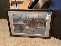 Framed and matted fine art print showing a snow-covered harbor village scene with boats and people under a cloudy sky.
