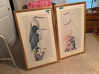Two large framed art prints by Art La May on carpeted floor. Prints feature herons and egrets with floral elements, gold wood frames.