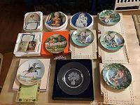 Lot of 11 collector plates arranged on a surface showing different designs including Tom Sawyer, Princess Diana, Balloon Man, Liberty Tree. Plates are in boxes or styrofoam holders.
