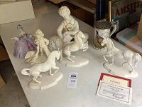 Photo showing seven porcelain collectibles on a table including three white horses, two boys with animals, a lady figurine in purple dress and a stein with painted game bird.