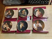 Six William A. Bouguereau collector plates on their original red boxes with Certificates of Authenticity, arranged on a table.