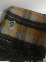 Folded blanket showing Ayres brand label sewn onto the fabric with a plaid pattern in yellow, grey, and black.