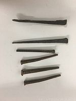 Top view of six antique square nails displayed on a white surface, showing varied lengths and rusted condition.