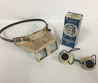 Vintage magnifying glasses with headband and vintage sun lamp goggles with original box on white background.