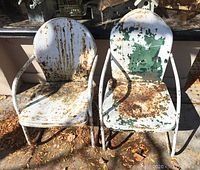 Pair of vintage motel metal patio chairs, rusty with peeling white and green paint, viewed frontally in outdoor setting with fallen leaves.