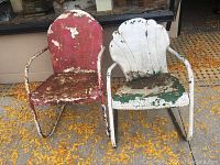 Front view of two vintage metal patio motel chairs with peeling paint and rust, one red, one white and green, on a leaf-covered surface.
