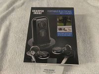 Front view of box showing Sharper Image Portable Electronic Key Finder with two black round key ring fobs attached to keys and a black tabletop cradle for remote transmitter storage.