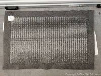 Gray rectangular rug with a patterned center and solid border, shown on a white and gray floor surface with tagging sticker visible