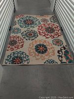 Rectangular rug laid out on concrete floor, showing full pattern with cream background and colorful Suzani floral designs.