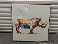Framed multicolor rhinoceros painting on a light background with artist signature and sticker tag on top center of frame.